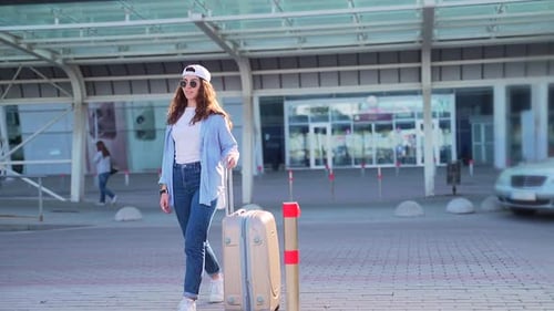 Cheerful Young Woman in Summer Cap and Sunglasses Walking Near Airport with Suitcase. Happy Lady