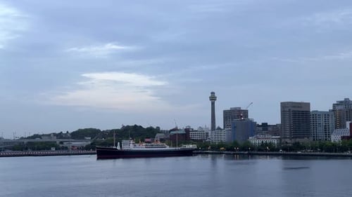 Dusk settles over city skyline with iconic tower and moored ship, gentle water in the foreground