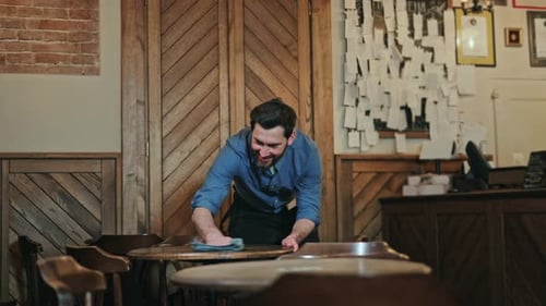 Cheerful Waiter Cleaning Table Surfaces with Wet Cloth at End of Working Day