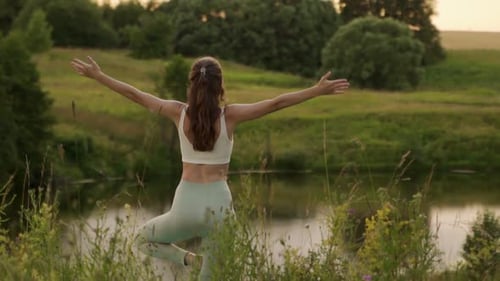 Young Woman Stretches Her Arms Up Doing Yoga Against the Background of a Pond