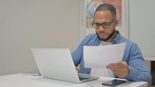 Man Reviews Document at Laptop in Bright Office