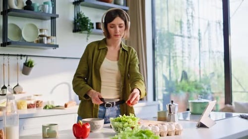 Joyful Brunette Listening Music in Earbuds Stirring Vegetables in Kitchen Area