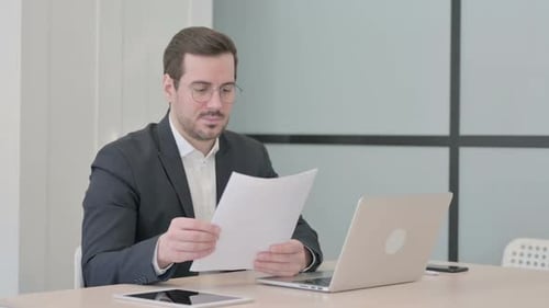 Man Reviews Documents While Using Laptop at Desk