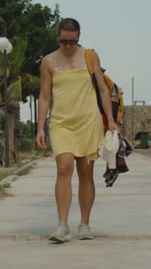 Vertical video. A Woman Walks Along a Path, Wrapped in a Yellow Towel After Swimming, With a Backpac