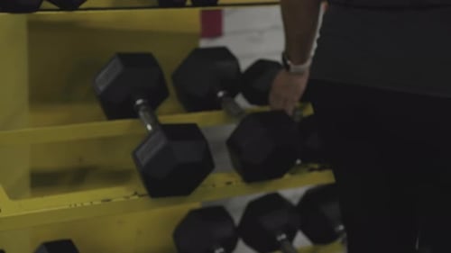 Woman in the gym approaches the dumbbell rack, picks up two weights. Slow-motion, close-up.