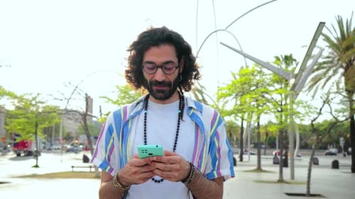 Young Adult Man Using His Smartphone Messaging and Chatting While Walking Through the Street