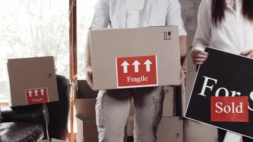 Couple Holding Moving Box and Sold Sign Indoors