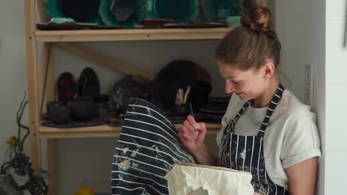 A female potter with a smile draws a ceramic plate with paint brush.