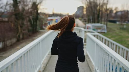 Young woman in black sports outfit running on the bridge in the city during day.