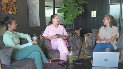 Diverse women hosting in lounge, right host leading, center reading tablet, microphone present