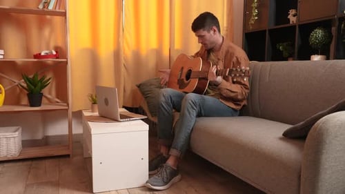 Man Plays Guitar While Looking at Laptop Indoors