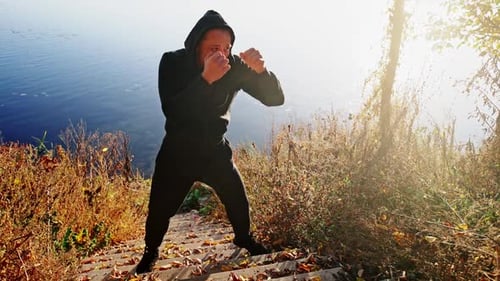 Young Active Man Trains in a Box Against the Background of a Lake in a Cool Autumn Morning