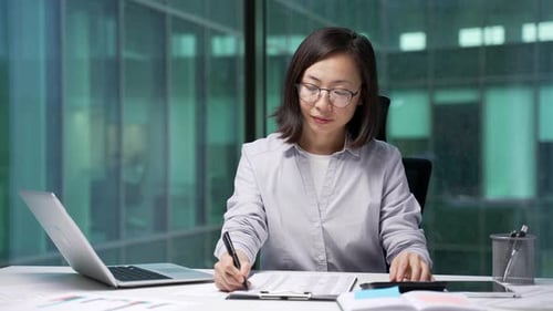 Asiatische Geschäftsfrau, die finanzielle Berechnungen mit einem Taschenrechner durchführt und am Schreibtisch am Arbeitsplatz sitzt