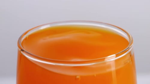 Close Up Of Ice Cube Falling In A Glass Of Orange Juice On The Light White Screen Background