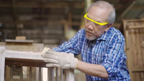 An elderly Asian male carpenter sanding a wooden piece of furniture in a carpentry workshop