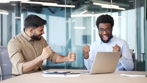 Two happy businessmen celebrating successful results using laptop computer at office desk.