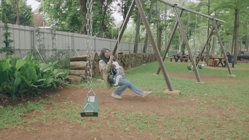 A Southeast Asian mother and daughter in hijab are playing on a swing in the park happily