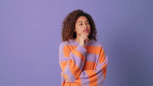 Young woman can't make decision looking at camera on lilac background in studio