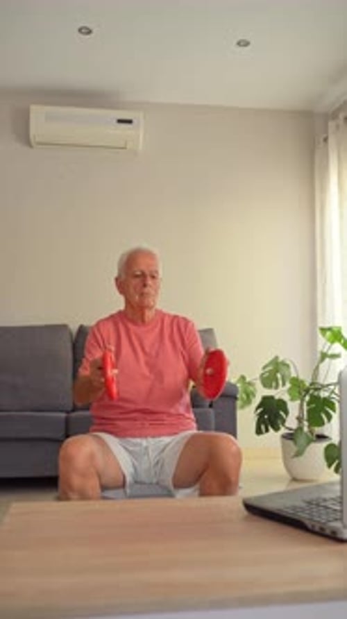 Senior man in sportswear doing dumbbell workout following online training on laptop at home