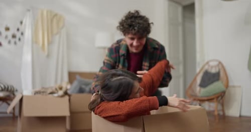 Cheerful Guy with Curly Hair Drags His Girlfriend in a Cardboard Box During a Hilarious Scene While