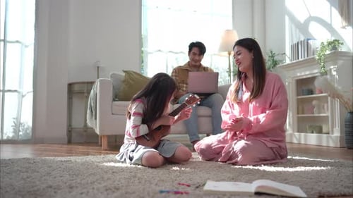 A young girl playing the ukulele for her mother while her father works on a laptop in the background