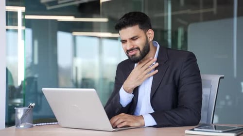 Bearded man works at desk, touches throat in pain