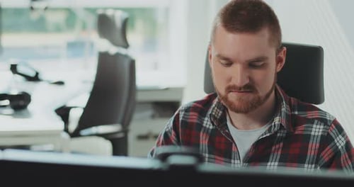 Ein junger bärtiger Mann sitzt an einem Computer, arbeitet im Büro, fühlt sich glücklich, zeitgenössisches Design, gedreht