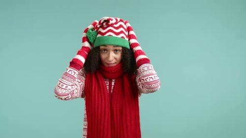 Süße Frau im Weihnachtspullover, Elfenmütze und rotem Schal, die kalt wird, wenn sie auf die Uhr schaut