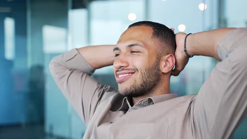 Young happy businessman relaxes with hands behind head while sitting at workplace in modern office.