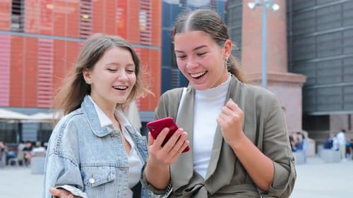 Two Young Women Cheering at Phone News in City