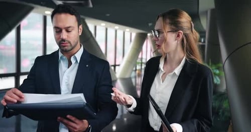 Two Colleagues Walk Through An Office Corridor, Man Reviews A Document, While The Woman Speaks. -