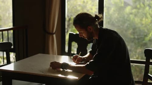 Man Writes at Table Near Window in Sunlight