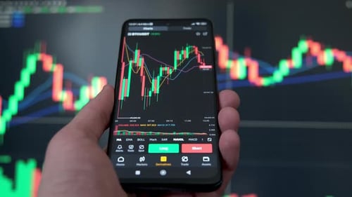 An Investor Holds a Smartphone in His Hand and Analyzes the Crypto Exchange Market In the Background