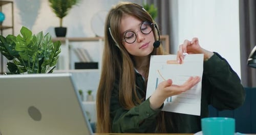 Business woman worker in glasses wears headset discussing report with chart during video conference
