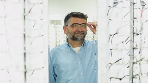 Apuesto hombre mayor barbudo escogiendo gafas nuevas en una tienda de óptica mirándose al espejo