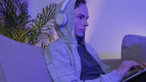 A Young Man Works Lying on the Couch with Headphones in the Evening