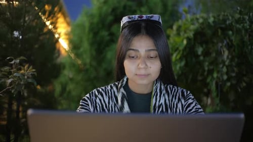 Uzbek Woman in Traditional Clothing and Headdress Works on a Laptop Outdoors