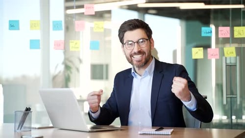 Joyful businessman celebrating successful achievement at workplace in office. Man with laptop