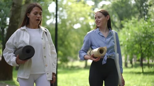 Two Women Carrying Yoga Mats and Walking Through a Sunlit Park