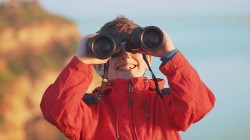 Happy boy looks through big binoculars into distance of sea against sunset
