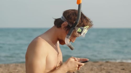 Un homme à la barbe rousse sur la plage, portant des lunettes de plongée et un tuba, regardant quelque chose sur son p