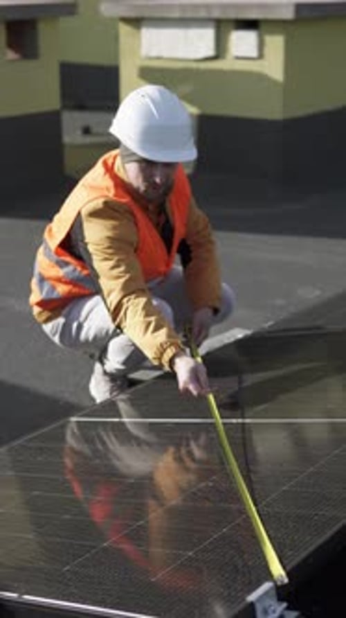 A construction worker in protective gear measures a solar panel on a roof during installation.
