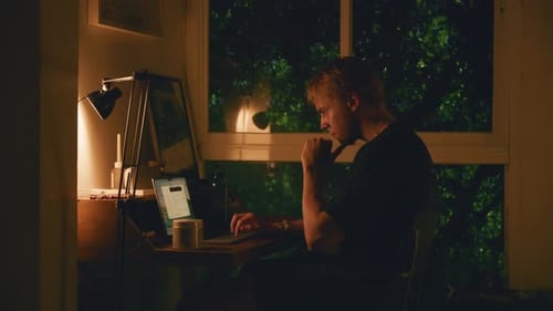 Young Student Studies Late at Night on His Laptop in a Dark Room Illuminated By a Desk Lamp While