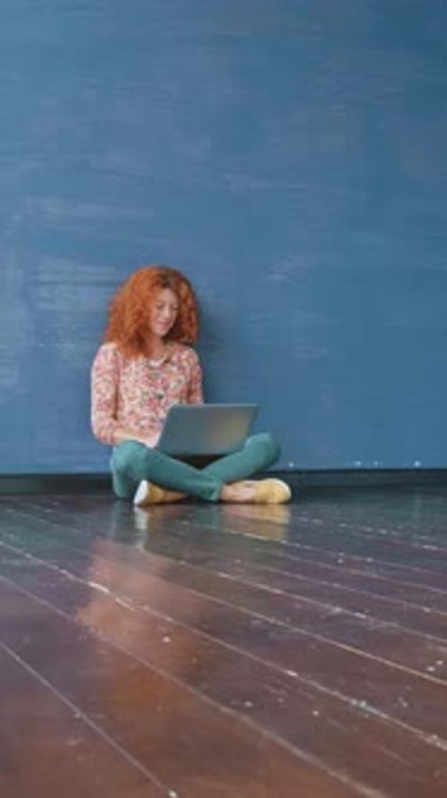 Young Woman with Curly Red Hair is Sitting on Wooden Floor Working on Laptop Against a Blue Wall
