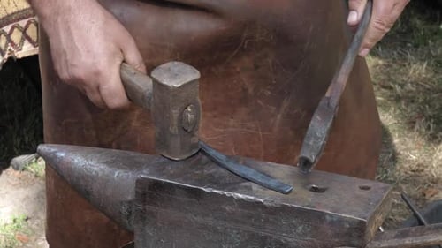Traditional blacksmith forges a piece of metal with a hammer. He works outside. Handheld.