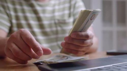 Young Man Counting US Dollar Bills At Table With Kitchen Background Close Up