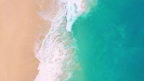 Aerial drone footage of massive turquoise waves crashing on a sandy tropical beach.