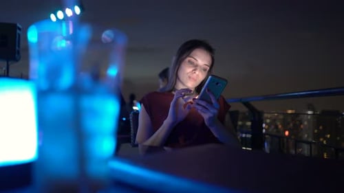 Young woman enjoying city nightlife browsing her smartphone on a rooftop terrace