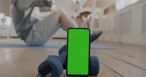 Closeup of Smartphone with Chroma Key Green Screen Near Dumbbells and Sportsman Doing Crunches at