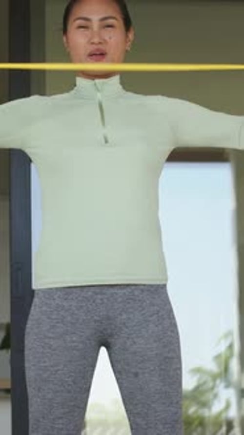 Vídeo vertical: puxando uma faixa amarela no peito, mulher asiática tonificando os ombros em casa com uma blusa verde